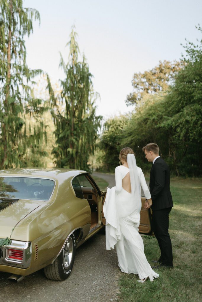 Bride and groom pose in front of their classic car, capturing a timeless moment on their wedding day